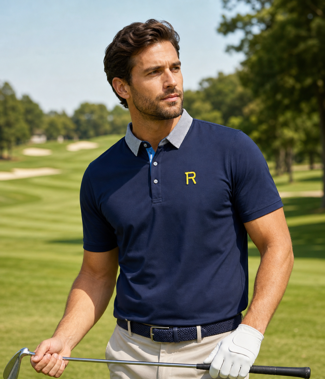 A man on a golf course wearing a navy blue short-sleeve polo shirt with a light gray collar, a yellow embroidered letter 'R' on the chest, beige pants, and a white golf glove, holding a golf club.